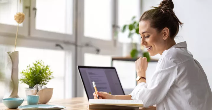 Shows a woman writing in her notepad next to her laptop