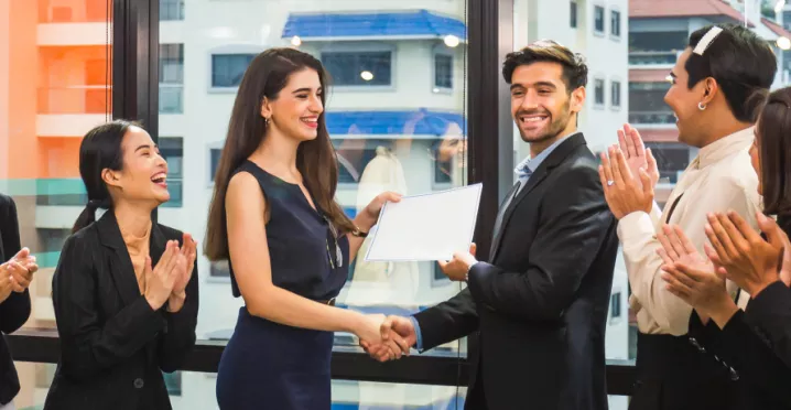 Image shows a coworker presenting an award to another coworker in front of a group of teammates.