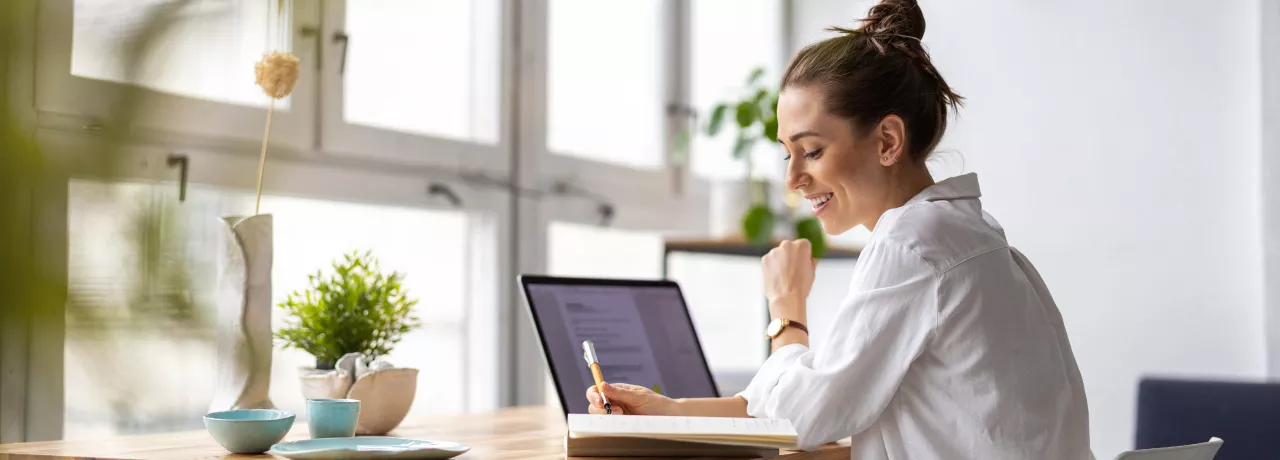 Shows a woman writing in her notepad next to her laptop