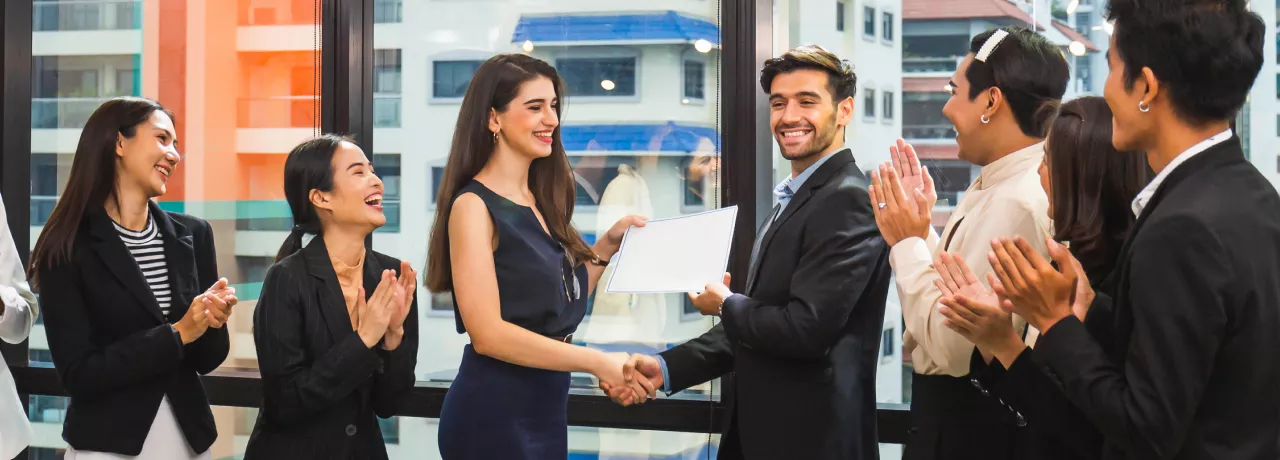 Image shows a coworker presenting an award to another coworker in front of a group of teammates.