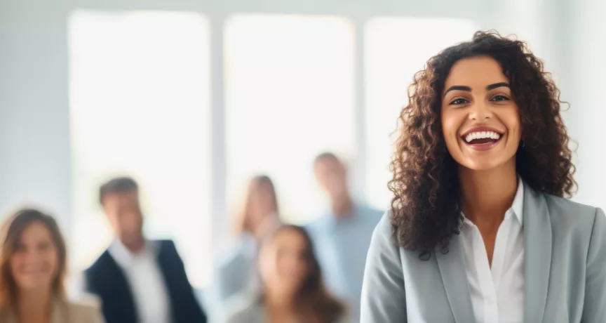 Shows a woman smiling at the camera with colleagues behind her