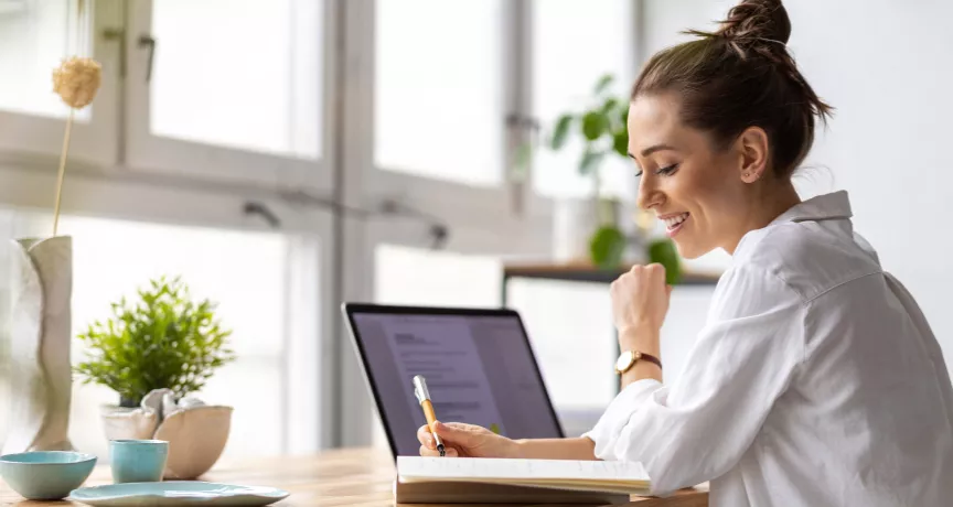Shows a woman writing in her notepad next to her laptop