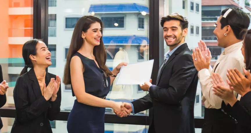 Image shows a coworker presenting an award to another coworker in front of a group of teammates.