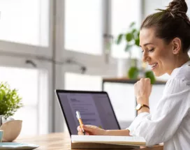 Shows a woman writing in her notepad next to her laptop