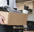 Image shows a person leaving an office with a box of items.